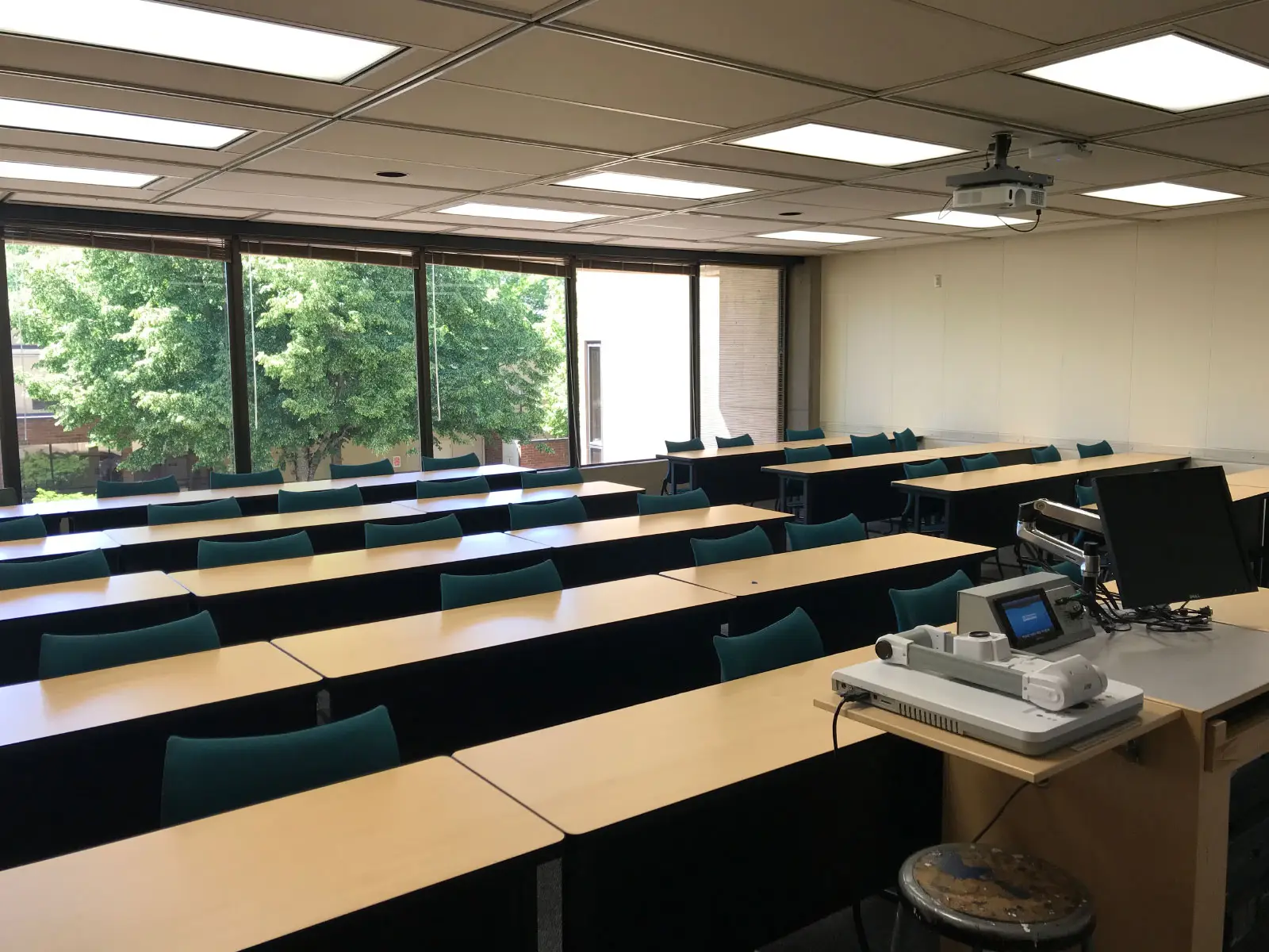 Rows of tables and blue chairs in front of a podium, next to ceiling-high windows in a classroom at the Oregon City campus